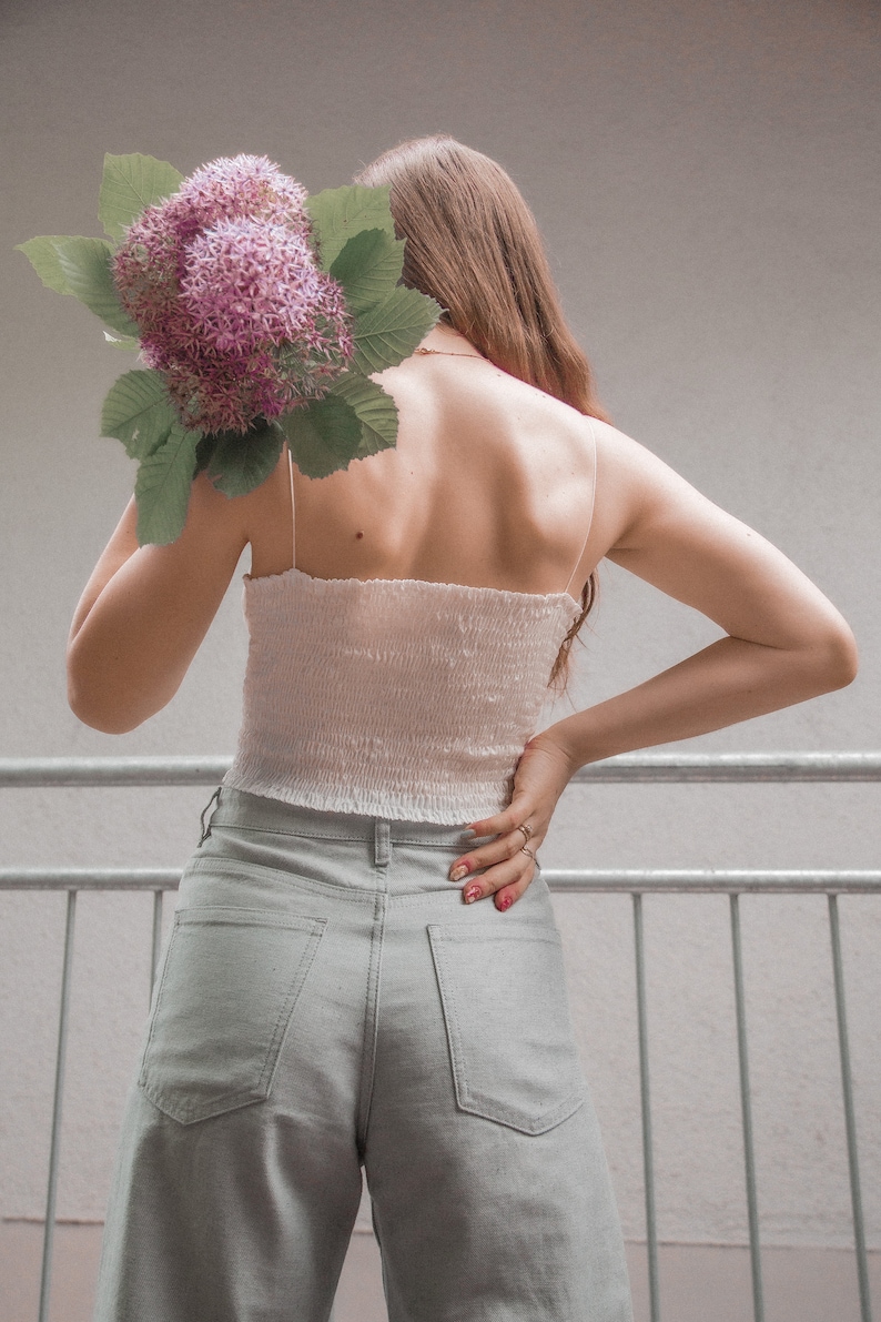 May include: A person wearing a white crop top and light gray wide-leg pants. They are holding a bouquet of pink flowers behind their back.