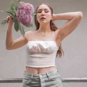 May include: A woman wearing a white crop top with a ruffled neckline and light green jeans. She is holding a bouquet of purple flowers.