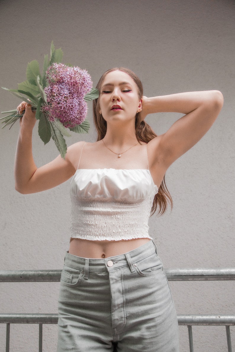 May include: A woman wearing a white crop top with a ruffled neckline and light green jeans. She is holding a bouquet of purple flowers.
