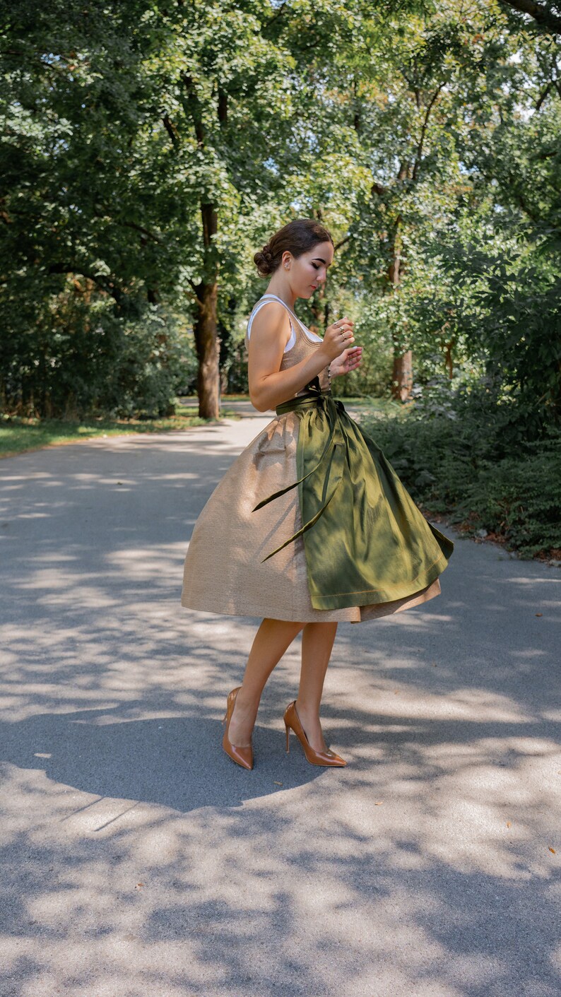 May include: A woman wearing a traditional dirndl dress with a brown bodice and a green apron. She is standing on a paved path in a park with trees in the background.