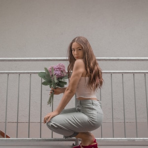 May include: A woman wearing a white crop top and light blue jeans sits on a concrete ledge with a bouquet of purple flowers. She is wearing red heeled shoes.
