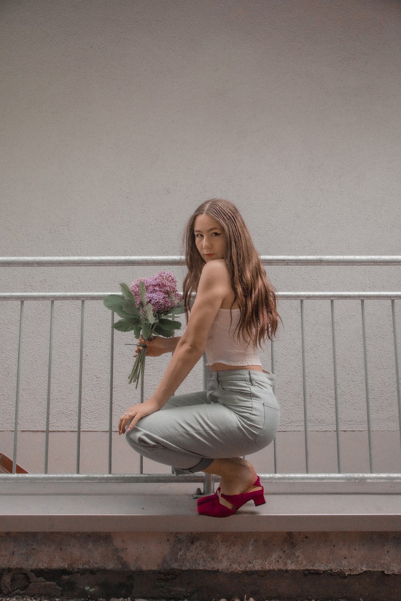 May include: A woman wearing a white crop top and light blue jeans sits on a concrete ledge with a bouquet of purple flowers. She is wearing red heeled shoes.