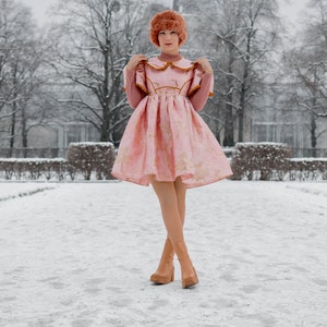 May include: A woman wearing a pink dress with gold embroidery and brown boots stands in a snowy park. The dress has a white collar and a ruffled neckline. The woman is looking at the camera.