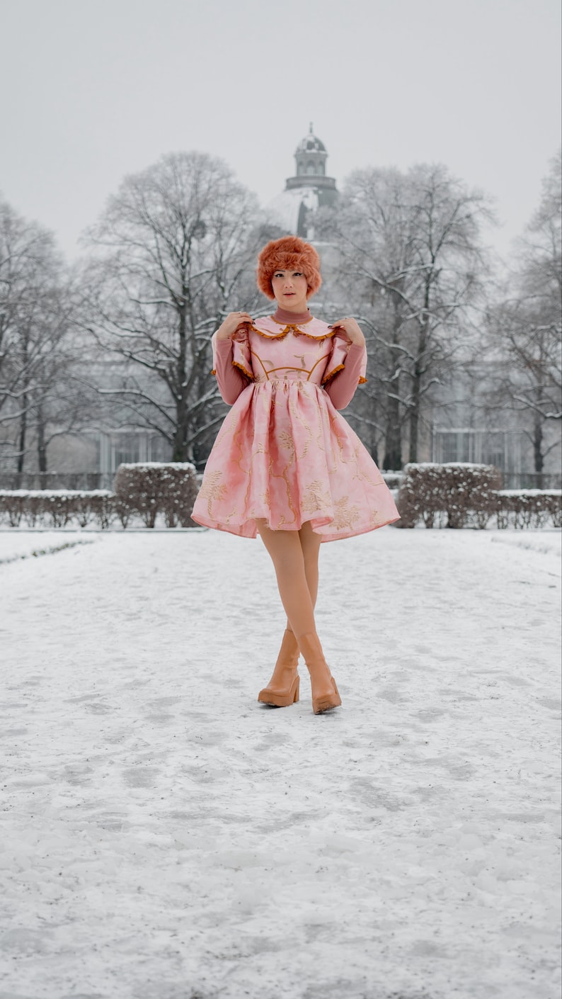 May include: A woman wearing a pink dress with gold embroidery and brown boots stands in a snowy park. The dress has a white collar and a ruffled neckline. The woman is looking at the camera.