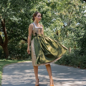 May include: A woman wearing a green and brown dirndl dress with a white blouse and brown heels. She is standing on a paved path in a park.