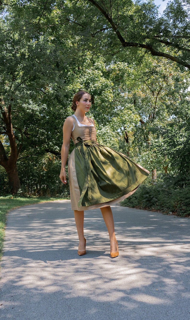 May include: A woman wearing a green and brown dirndl dress with a white blouse and brown heels. She is standing on a paved path in a park.