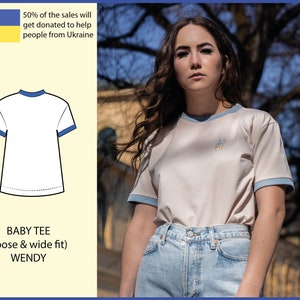 Könnte beinhalten: Ein beiges Baby-T-Shirt mit blauen Abschlüssen und einem kleinen gestickten Design. Das T-Shirt ist locker und weit geschnitten. 50% des Erlöses werden gespendet, um Menschen aus der Ukraine zu helfen.