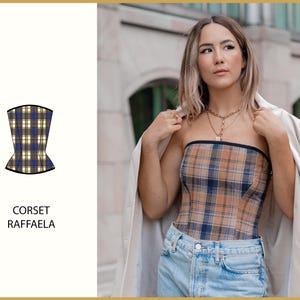 May include: A plaid corset with a black trim. The corset is strapless and has a fitted silhouette. The model is wearing the corset with a white blazer and blue jeans.