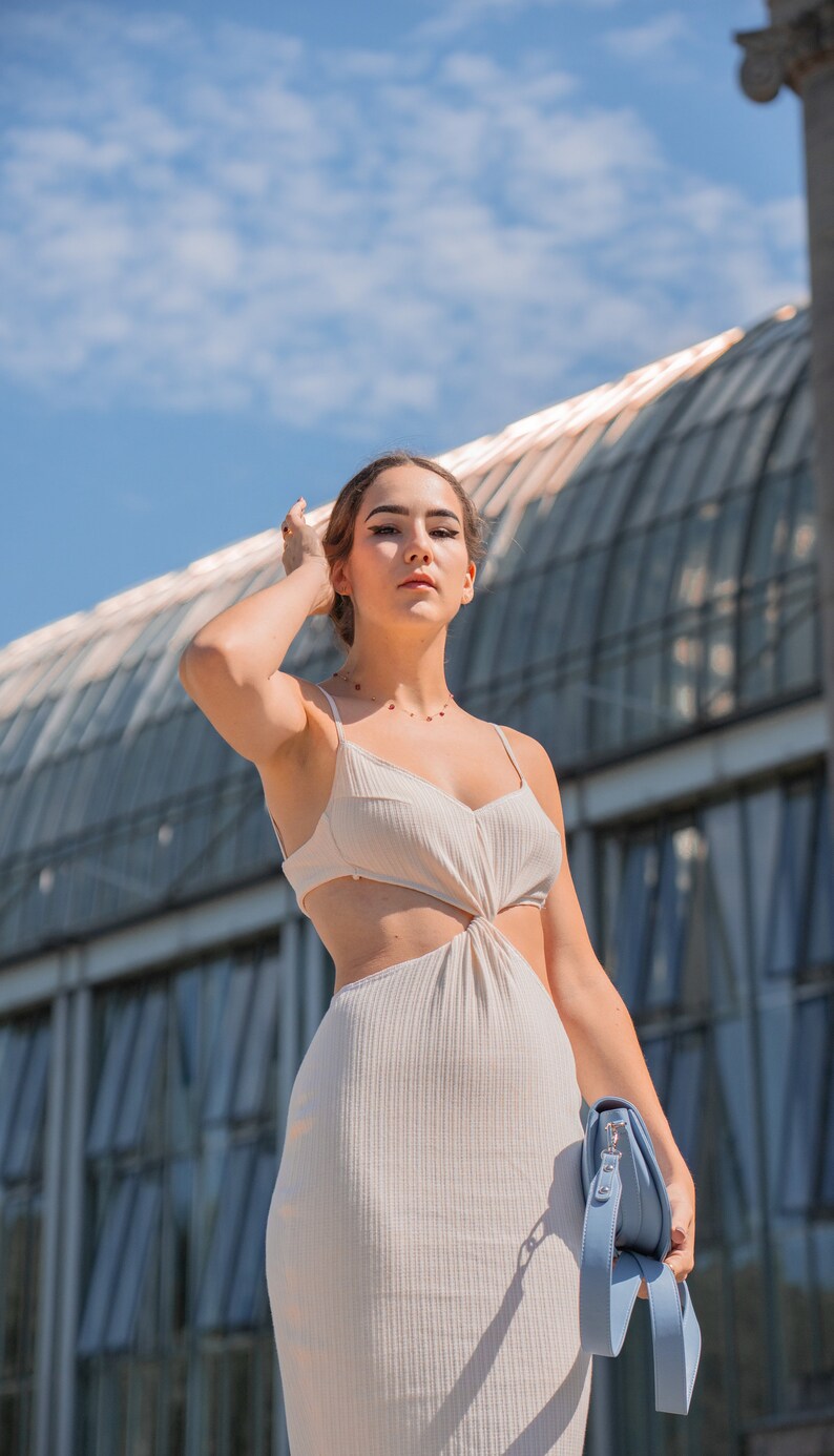 Pu&ograve; includere: Una donna indossa un abito bianco con scollatura e un dettaglio annodato in vita. Tiene una borsa a mano blu chiaro con una lunga tracolla.