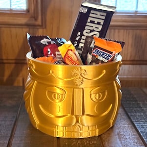 May include: A gold-colored nutcracker-shaped container filled with assorted candy bars including Hershey's milk chocolate, Snickers, and Starburst. The container features a molded face with a crown, mustache, and teeth, serving as a bowl for sweets.