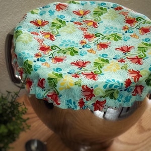 May include: A light blue bowl cover with a floral pattern of red, yellow, and green flowers and leaves. The cover is placed over a stainless steel bowl, creating a decorative and functional kitchen accessory.