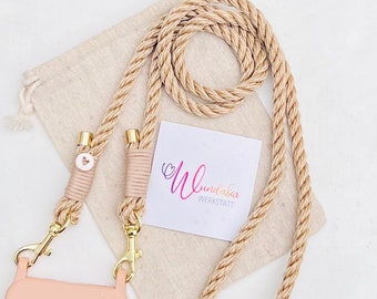 May include: A beige rope phone strap with gold clasps and a small pink heart charm. The strap is coiled and laid on a beige fabric pouch with the text "Wunderbar Werkstatt" printed on a white card.