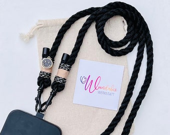 May include: Black phone strap with a smiley face charm. The strap is made of thick rope and has a black metal clasp. The strap is lying on a beige fabric bag with a white tag that says "wundabar werkstatt".