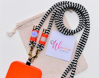 May include: A black and white patterned phone strap with a bright orange phone case and a gold clasp. The strap has a small smiley face charm with a purple face.