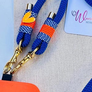 May include: A blue and orange phone strap with gold clasps and a heart-shaped charm. The strap is made of woven rope and has a white and blue patterned design. The text "Wunderbar Werkstatt" is visible in the background.