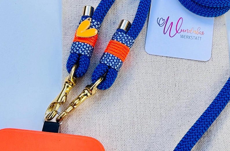 May include: A blue and orange phone strap with gold clasps and a heart-shaped charm. The strap is made of woven rope and has a white and blue patterned design. The text "Wunderbar Werkstatt" is visible in the background.