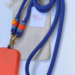 May include: A blue phone strap with orange and white accents and gold clasps. The strap is coiled and next to a small white bag with the text "Wunderbar Werkstatt".