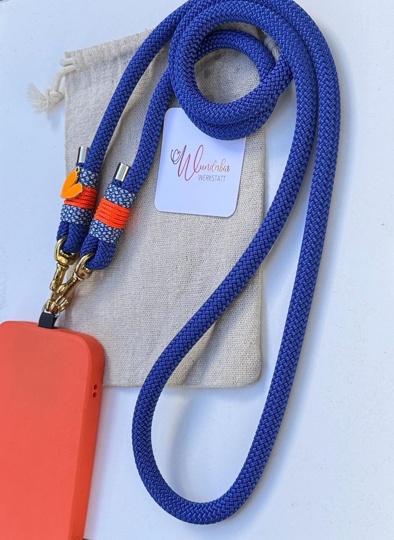 May include: A blue phone strap with orange and white accents and gold clasps. The strap is coiled and next to a small white bag with the text "Wunderbar Werkstatt".