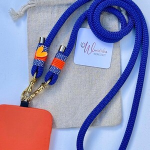 May include: A blue phone lanyard with a gold clasp and orange and white accents. The lanyard has a small heart charm and is attached to a bright orange phone case. The lanyard is on a beige fabric background with a white tag that says "Wundabar Werkstatt".