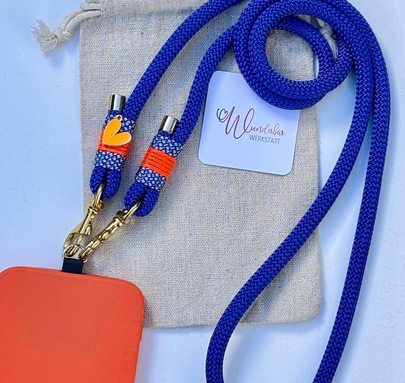 May include: A blue phone lanyard with a gold clasp and orange and white accents. The lanyard has a small heart charm and is attached to a bright orange phone case. The lanyard is on a beige fabric background with a white tag that says "Wundabar Werkstatt".