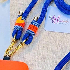 May include: A blue rope phone strap with orange and white accents. The strap has a gold metal clasp and a small orange heart charm. The strap is lying on a beige fabric surface. The text "Wundabar Werkstatt" is visible on a white card.