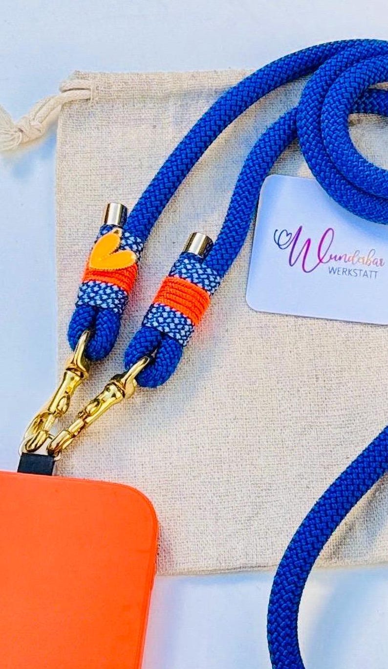 May include: A blue rope phone strap with orange and white accents. The strap has a gold metal clasp and a small orange heart charm. The strap is lying on a beige fabric surface. The text "Wundabar Werkstatt" is visible on a white card.