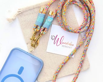 May include: A multicolored braided rope phone lanyard with gold-toned metal clasps and light blue cord accents. A small heart-shaped charm with an eye design is attached. A light blue phone case and a white card with the text Wundabar WERKSTATT are present.