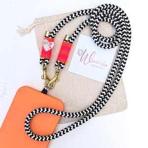 Peut inclure: Une lanière de téléphone à motif chevron noir et blanc avec des fermoirs dorés et des accents rose fluo. La lanière est attachée à une coque de téléphone orange. Un petit sac et une carte avec le texte "Wundabar Werkstatt" sont en arrière-plan.