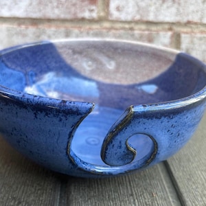 May include: A blue ceramic yarn bowl with a swirl-shaped yarn guide. The bowl has a glossy finish and a two-tone design, with a lighter blue interior and a darker blue exterior. The bowl is designed for holding yarn while knitting or crocheting.