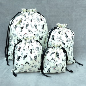 May include: Four white drawstring bags with black trim and a black and white cat print. The bags are arranged in a pyramid shape, with the largest bag at the bottom and the smallest bag at the top.