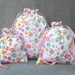 May include: Three drawstring bags with pink ribbon ties. The bags are made of white fabric with a colourful Easter egg pattern.