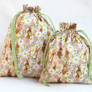 May include: Two drawstring bags with a floral print featuring brown bunnies and green leaves. The bags are made of fabric and have green ribbon ties.