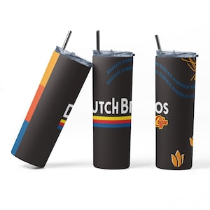 Op de afbeelding: Drie zwarte geïsoleerde bekers met zilveren rietjes. Eén beker heeft een kleurrijk geometrisch ontwerp, een andere heeft de tekst "Dutch Bros" met een rode, gele en blauwe streep, en de derde heeft de tekst "Dutch Bros Coffee" en een bloemenontwerp.