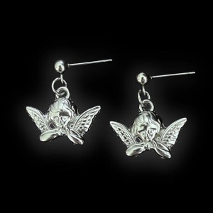 Peut inclure: Une paire de boucles d'oreilles en argent en forme d'ange avec un petit ange d&eacute;taill&eacute;. Les anges sont tourn&eacute;s vers l'avant, les ailes d&eacute;ploy&eacute;es. Les boucles d'oreilles sont sur un fond noir.