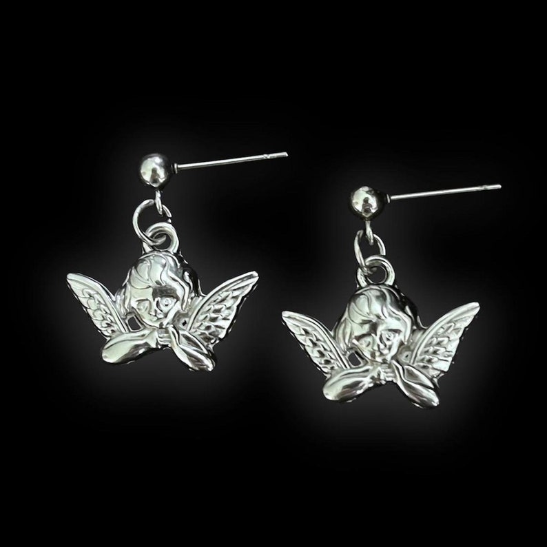 Peut inclure: Une paire de boucles d'oreilles en argent en forme d'ange avec un petit ange d&eacute;taill&eacute;. Les anges sont tourn&eacute;s vers l'avant, les ailes d&eacute;ploy&eacute;es. Les boucles d'oreilles sont sur un fond noir.