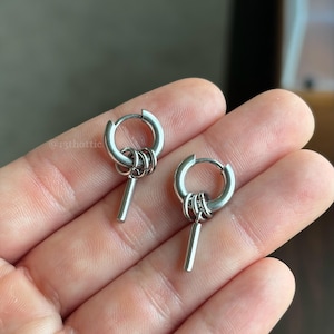 May include: A pair of silver-toned hoop earrings featuring a small dangling cylindrical bar charm attached to the hoop by three interconnected jump rings. The text @13thattic is visible in the upper left corner of the image. The earrings are metallic.