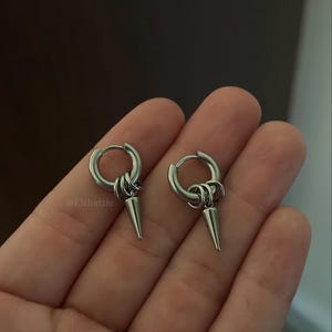 May include: A pair of silver-toned hoop earrings, each featuring a small circular hoop with two attached jump rings and a dangling metal spike charm. The text @13thattic is visible on the left earring. These earrings have a polished finish.