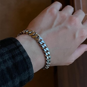 May include: A gold-colored curb chain bracelet. The bracelet is made of interlocking links and is worn on a wrist. The image shows the bracelet in detail, highlighting its texture and shine. The bracelet is a piece of jewelry.