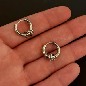 May include: A pair of silver-toned hoop earrings, each featuring a hinged closure and three small, loose rings attached to the main hoop. The metal has a polished, reflective finish. The text @13thallie is visible in the background area.