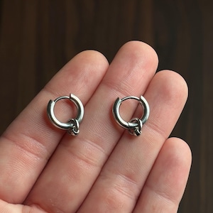 May include: A pair of small, polished silver-toned hoop earrings. Each circular hoop features a hinged closure mechanism and a single, small, spherical charm dangling from the bottom of the ring, creating a simple and clean metallic appearance.