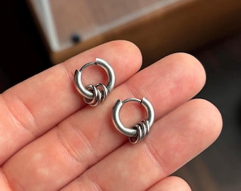 May include: A pair of silver-toned hoop earrings, each featuring a smooth circular band with three smaller, interconnected silver rings dangling from the bottom. The metal has a polished finish with subtle darkened detailing in the crevices.