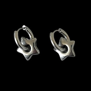 May include: A pair of silver-toned hoop earrings, each featuring a polished, five-pointed star charm suspended from the circular band. The metal has a smooth, reflective finish, and the design includes a hinged closure mechanism on the hoops.
