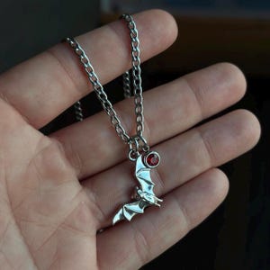 May include: A silver-toned metal necklace featuring a bat-shaped pendant with outstretched wings. A small, circular, deep red gemstone is set in a silver bezel attached to the top of the bat charm, which hangs from a standard silver-colored link chain.