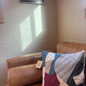 Peut inclure: Un coussin patchwork avec une variété de tissus bleus, rouges et beiges repose sur un canapé en cuir marron. Un coussin de soutien assorti est également visible. Une peinture de paysage encadrée est accrochée au mur au-dessus du canapé.