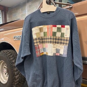 May include: A blue sweatshirt featuring a patchwork design on the front. The patchwork is made up of different coloured fabrics, including red, green, yellow, and blue.