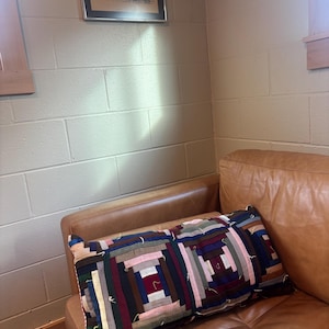 May include: A rectangular patchwork cushion with a log cabin design sits on a brown leather sofa. The cushion features various fabric colours, including blue, pink, and burgundy. A framed landscape painting hangs on the wall above the sofa.