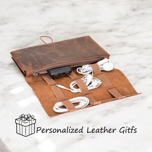 Puede incluir: Un rollo de viaje de cuero marr&oacute;n con m&uacute;ltiples correas el&aacute;sticas para sujetar cables y auriculares. El rollo est&aacute; abierto y tiene un cargador de tel&eacute;fono negro y auriculares blancos en su interior. "Personalized Leather Gifts" est&aacute; escrito en blanco en la parte inferior de la imagen.