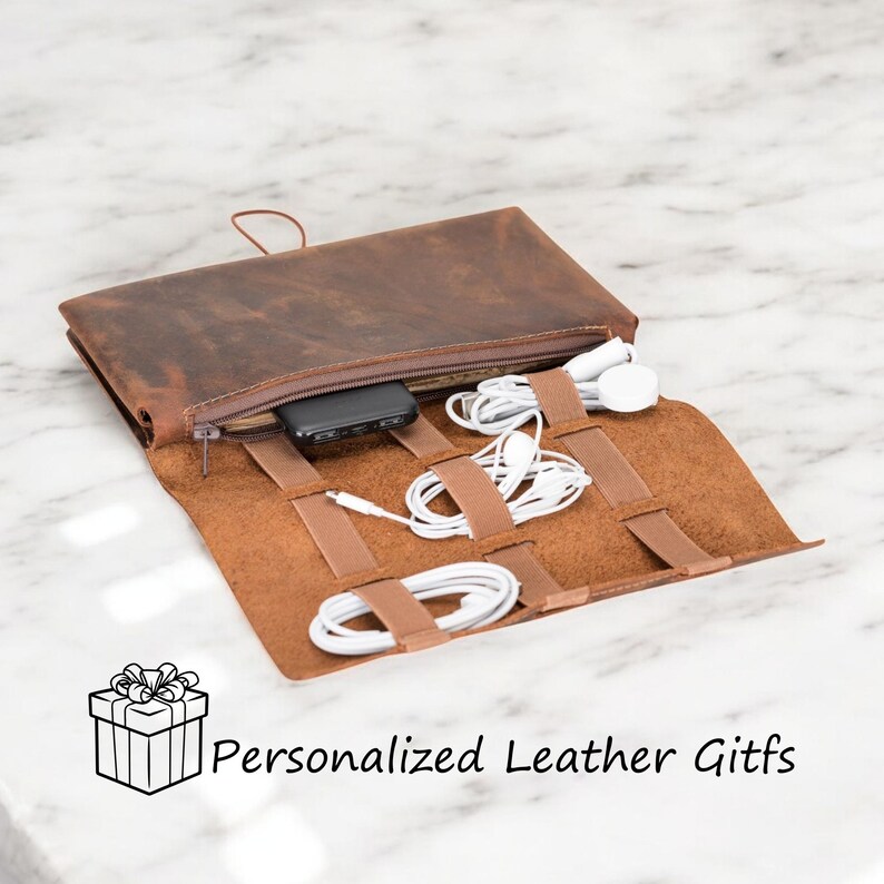 Puede incluir: Un rollo de viaje de cuero marr&oacute;n con m&uacute;ltiples correas el&aacute;sticas para sujetar cables y auriculares. El rollo est&aacute; abierto y tiene un cargador de tel&eacute;fono negro y auriculares blancos en su interior. "Personalized Leather Gifts" est&aacute; escrito en blanco en la parte inferior de la imagen.