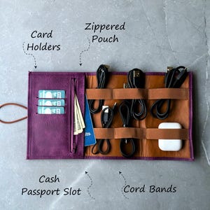 Puede incluir: Organizador de viaje de cuero morado con múltiples compartimentos para cables, efectivo, tarjetas y un pasaporte. El organizador tiene un bolsillo con cremallera, bandas elásticas para cables y una ranura para efectivo y pasaporte.