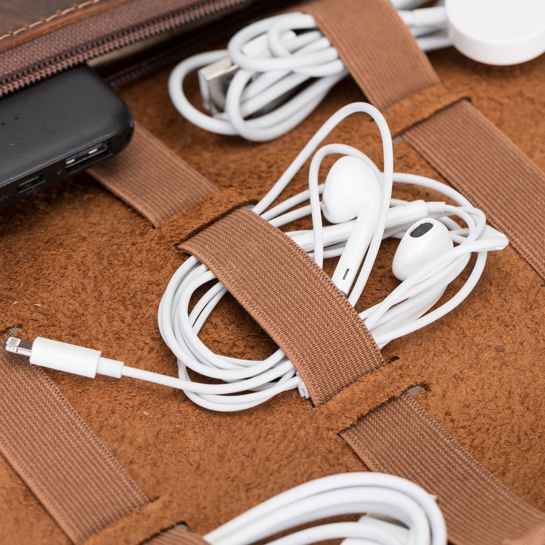 Puede incluir: Una funda de viaje de cuero marr&oacute;n con m&uacute;ltiples correas el&aacute;sticas para sujetar varios cables y accesorios electr&oacute;nicos. La funda est&aacute; abierta y muestra cables y auriculares blancos.
